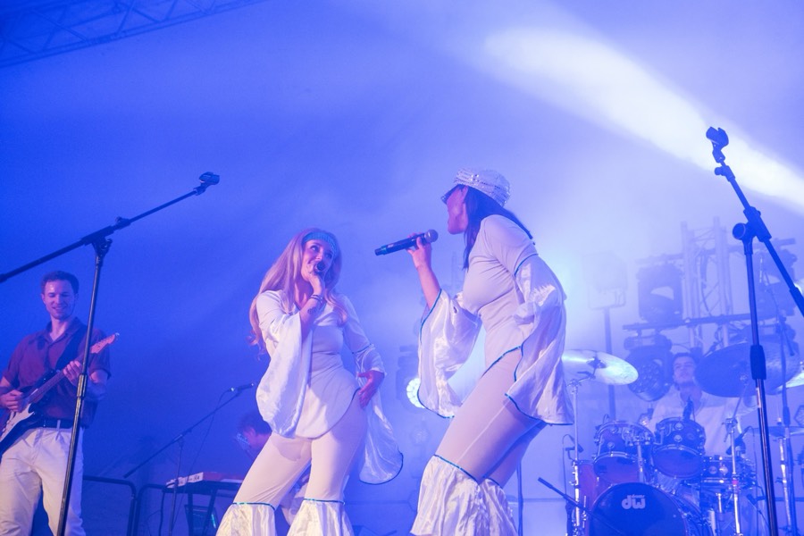 Two singers at the front of the stage in white costumes with guitar and drums behind them